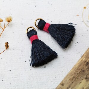 Op de afbeelding: Twee donkerblauwe kwastcharms met een roze accent en gouden springringen. De kwasten zijn gemaakt van zacht garen en zijn ongeveer 2,5 cm lang.