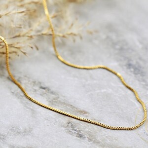May include: A gold chain necklace with a box chain style. The chain is lying on a light grey surface.