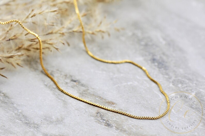 May include: A gold chain necklace with a box chain style. The chain is lying on a light grey surface.