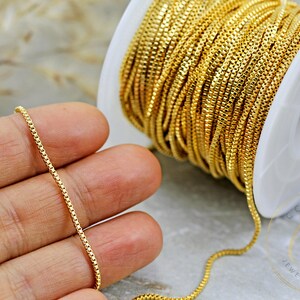 May include: A spool of gold-coloured chain, with a close-up of a section of the chain on a hand. The chain is a fine, box chain style.