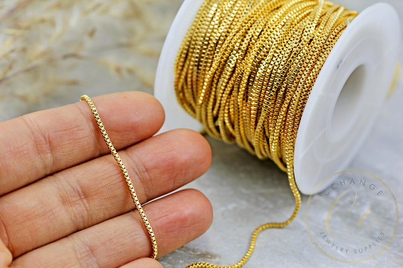 May include: A spool of gold-coloured chain, with a close-up of a section of the chain on a hand. The chain is a fine, box chain style.