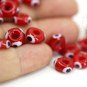Puede incluir: Primer plano de cuentas de ojo turco rojas y blancas. Las cuentas son redondas y tienen un agujero en el centro. Están esparcidas sobre una mano.