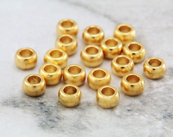 Könnte beinhalten: Eine Gruppe von 18 goldfarbenen Metallperlen. Die Perlen sind rund und haben ein Loch in der Mitte. Sie liegen lose auf einer weißen Marmorfläche.