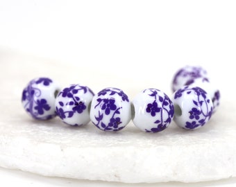 Puede incluir: Un conjunto de seis cuentas de cerámica blanca con un diseño floral morado. Las cuentas son redondas y tienen un acabado liso.