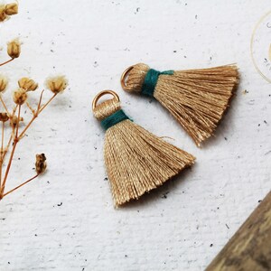 Op de afbeelding: Twee goudkleurige kwasten met teal draadaccenten en gouden ringen. De kwasten zijn gemaakt van fijne draden, tegen een getextureerde witte achtergrond met gedroogde bloemen.