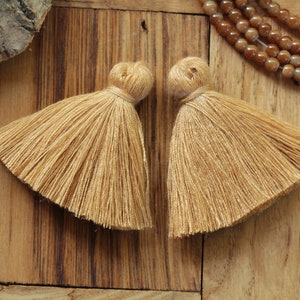 May include: Two beige tassel trims with a knotted top. The tassels are made of soft, fluffy fibers and are approximately 2 inches in length.