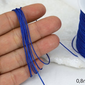Puede incluir: Un carrete de cordón elástico azul real, de 0,8 mm de grosor, para la elaboración de joyas o manualidades. El cordón se muestra enrollado alrededor de un carrete blanco y sostenido en una mano.