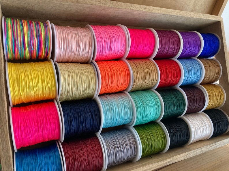 May include: A collection of spools of colourful waxed cotton cord, arranged in a wooden box. The cords are in various colours, including red, blue, green, yellow, pink, and purple.