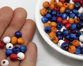 May include: A close-up of a hand holding a variety of wooden beads in blue, orange, red, and white. The beads are round and have a hole in the center. There are more beads in a white bowl in the background.