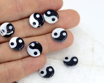 May include: A collection of small, round, black and white Yin and Yang symbol beads. The beads are glossy and have a smooth surface. The image shows several beads, some in focus and others slightly blurred, against a white background.