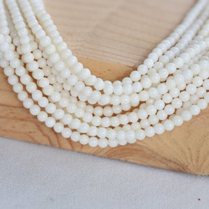 May include: A strand of white round shell beads, arranged in a fan shape on a wooden surface.
