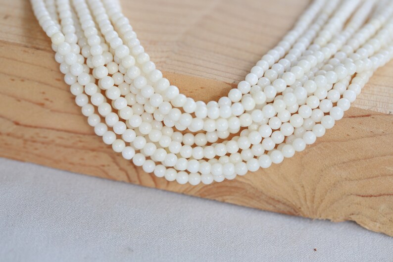 May include: A strand of white round shell beads, arranged in a fan shape on a wooden surface.