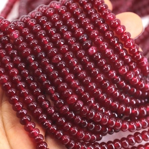 Puede incluir: Primer plano de hebras de pequeñas cuentas redondas de color rojo oscuro. Las cuentas están fuertemente ensartadas, creando una apariencia densa y texturizada. Las cuentas son de un color rojo oscuro intenso, con un acabado brillante. Las cuentas se sostienen en una mano.