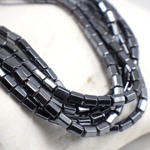 May include: A strand of black hematite beads, each bead is a square with slightly rounded edges.