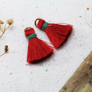 Op de afbeelding: Twee rode kwastbedels met gouden springringen en turquoise trim. De kwasten zijn gemaakt van zacht, pluizig garen en zijn perfect om een ​​bohemien touch toe te voegen aan sieraden of knutselwerk.