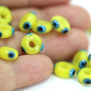 Puede incluir: Primer plano de cuentas de vidrio amarillas con diseños de ojo turco azul y negro. Las cuentas tienen un agujero en el centro para enhebrar.