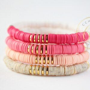 May include: Four stacked bracelets with pink, peach, and white ceramic beads and gold spacers. The bracelets are layered in a gradient from pink to white.