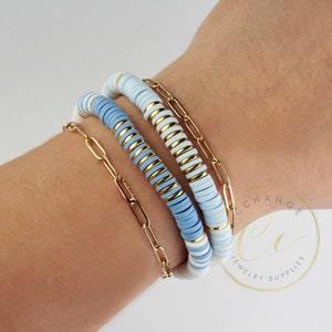 May include: A stack of three bracelets with blue and white ceramic beads and a gold chain link bracelet. The bracelets are on a person's wrist.