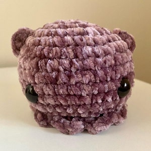May include: A plush purple crocheted octopus with black eyes. The octopus is made of a soft, fuzzy material.