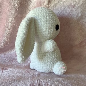 May include: A white crocheted bunny rabbit plush toy with black eyes. The bunny is sitting on a pink fuzzy surface.