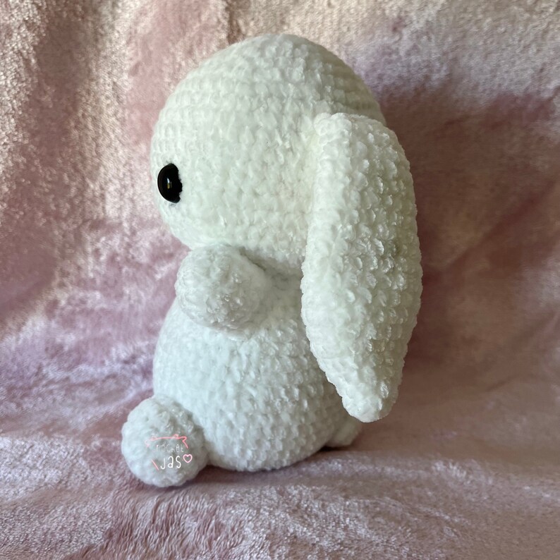 May include: White crocheted bunny rabbit plush toy with black eyes. The bunny is sitting on a pink fuzzy surface.