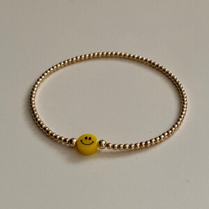 Puede incluir: Un brazalete de cuentas doradas con un dije de carita sonriente amarilla.