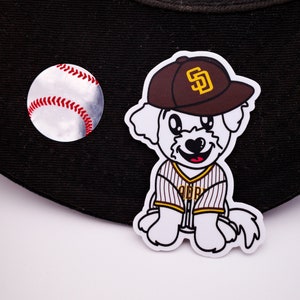 May include: A white dog wearing a brown baseball cap with a gold "SD" logo and a brown and white striped baseball jersey. The dog is sitting on a black baseball cap with a white baseball on the side.