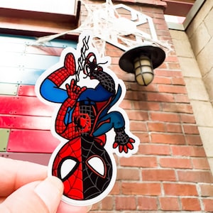 May include: A red and black cartoon superhero character hangs upside down from a web. The character is wearing a blue and red suit with a spider logo on the chest. The background is a brick wall.