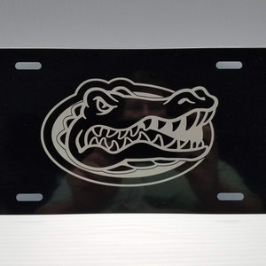 May include: Black license plate featuring a silver outline of an alligator head with sharp teeth. The alligator is inside an oval shape. The plate has four mounting holes.