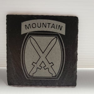 May include: Black slate coaster with a white engraved image of a crossed sword design and the word "MOUNTAIN" above it.