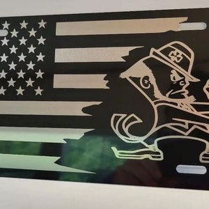 May include: A black metal license plate featuring a silver and black American flag with a white leprechaun figure in a green shamrock hat. The leprechaun is holding a club and has a beard.