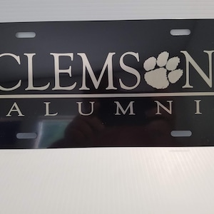 May include: Black metal license plate with white text that reads "CLEMSON ALUMNI" and a paw print.