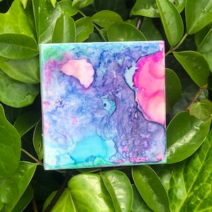 May include: Abstract art painting on a square tile with shades of blue, pink, and green. The colours are blended together in a swirling pattern.