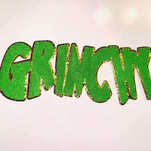 May include: Green glitter word "GRINCH" with gold glitter trim, set against a white background. The letters are bold and stylized, with a textured, sparkling appearance. The overall design is festive and eye-catching.