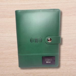 May include: A dark green leather notebook with a black label that says "XHF 1932". The notebook has a silver button and a wireless charging symbol embossed on the front.
