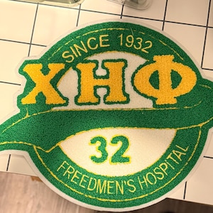 May include: A circular tufted rug featuring a green and white background with yellow Greek letters XHΦ. The text reads SINCE 1932, 32, and FREEDMEN'S HOSPITAL. The design includes a prominent green swoosh across the center, creating a textured emblem.