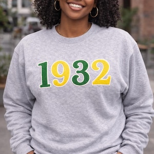 May include: A heather gray sweatshirt with the number "1932" in green and yellow lettering. The sweatshirt is long-sleeved and has a crew neck. The person is wearing gold hoop earrings.