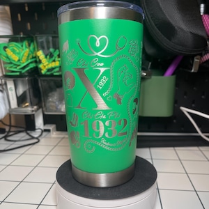 May include: Green stainless steel tumbler with a silver engraved design. The design includes a large letter X, the words "Chi Omega" and "Chi Eta Phi", and the years "1932" and "1982".