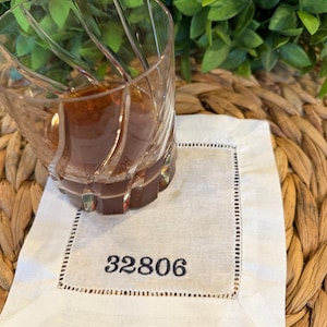 May include: A clear, patterned glass filled with a brown liquid sits on a white linen coaster. The coaster has a stitched border and the number "32806" printed in black.