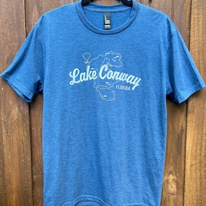 May include: Blue t-shirt with a crew neck and short sleeves. The shirt features a white graphic of Lake Conway, Florida, with the text "Lake Conway Florida" in a script font. The shirt is hanging on a black hanger against a wooden background.