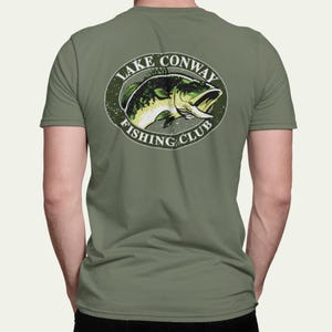 May include: Olive green t-shirt featuring a circular graphic of a fish with the text "Lake Conway Fishing Club". The fish is green and yellow, with the text in white. The shirt is a short-sleeved crew neck.