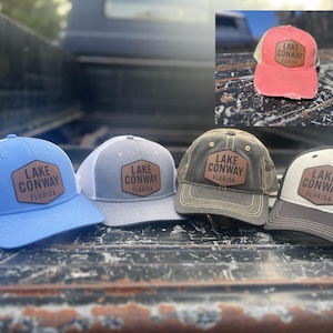 May include: Five different trucker hats with a brown leather patch that says "Lake Conway, Florida". The hats are in various colors, including blue, gray, black, and camouflage.
