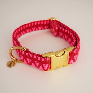 May include: A pink and gold dog collar with a geometric pattern. The collar has a gold buckle and a gold D-ring.
