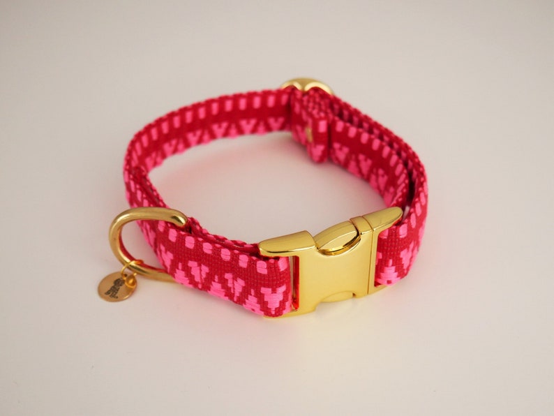 May include: A pink and gold dog collar with a geometric pattern. The collar has a gold buckle and a gold D-ring.