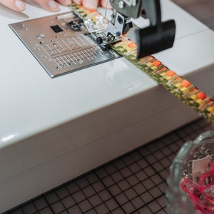 May include: A close-up of a sewing machine with a colorful fabric being sewn. The machine has a white body and a black foot pedal. The fabric is a mix of yellow, green, and orange.