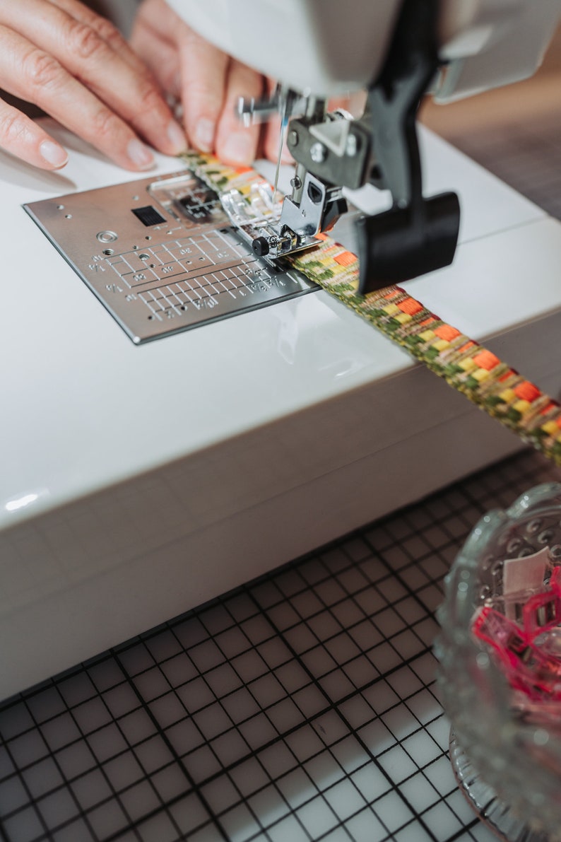 May include: A close-up of a sewing machine with a colorful fabric being sewn. The machine has a white body and a black foot pedal. The fabric is a mix of yellow, green, and orange.