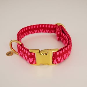May include: A pink dog collar with a gold buckle and a pink and white geometric pattern. The collar has a small gold tag with the word "handmade" on it.