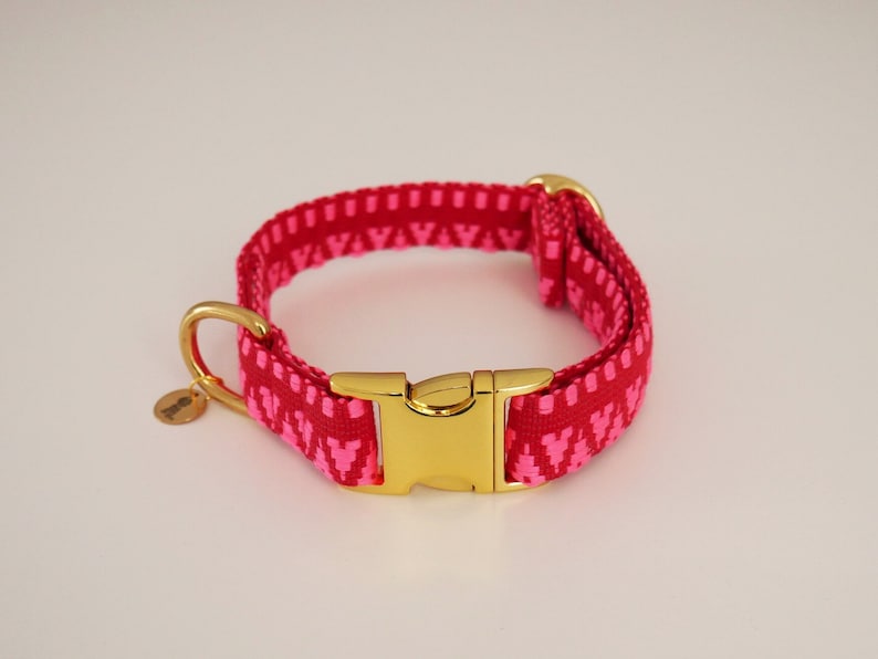 May include: A pink dog collar with a gold buckle and a pink and white geometric pattern. The collar has a small gold tag with the word "handmade" on it.
