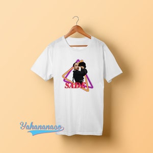 May include: White t-shirt with a graphic of Sade in a black outfit with a pink and yellow triangle design and the word "SADE" in pink.