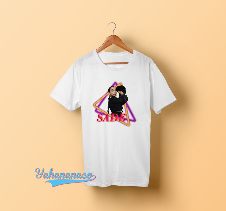 May include: White t-shirt with a graphic of Sade in a black outfit with a pink and yellow triangle design and the word "SADE" in pink.